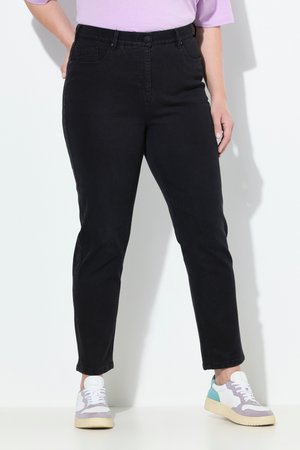 Person iført sorte slim-fit bukser, hvide og turkise sneakers og en lyse lilla skjorte, stående mod en lys baggrund.