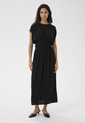 Mujer con cabello largo y rizado, vestida con un vestido midi negro suelto con cintura fruncida y mules negras de tacón kitten, posando sobre un fondo blanco.