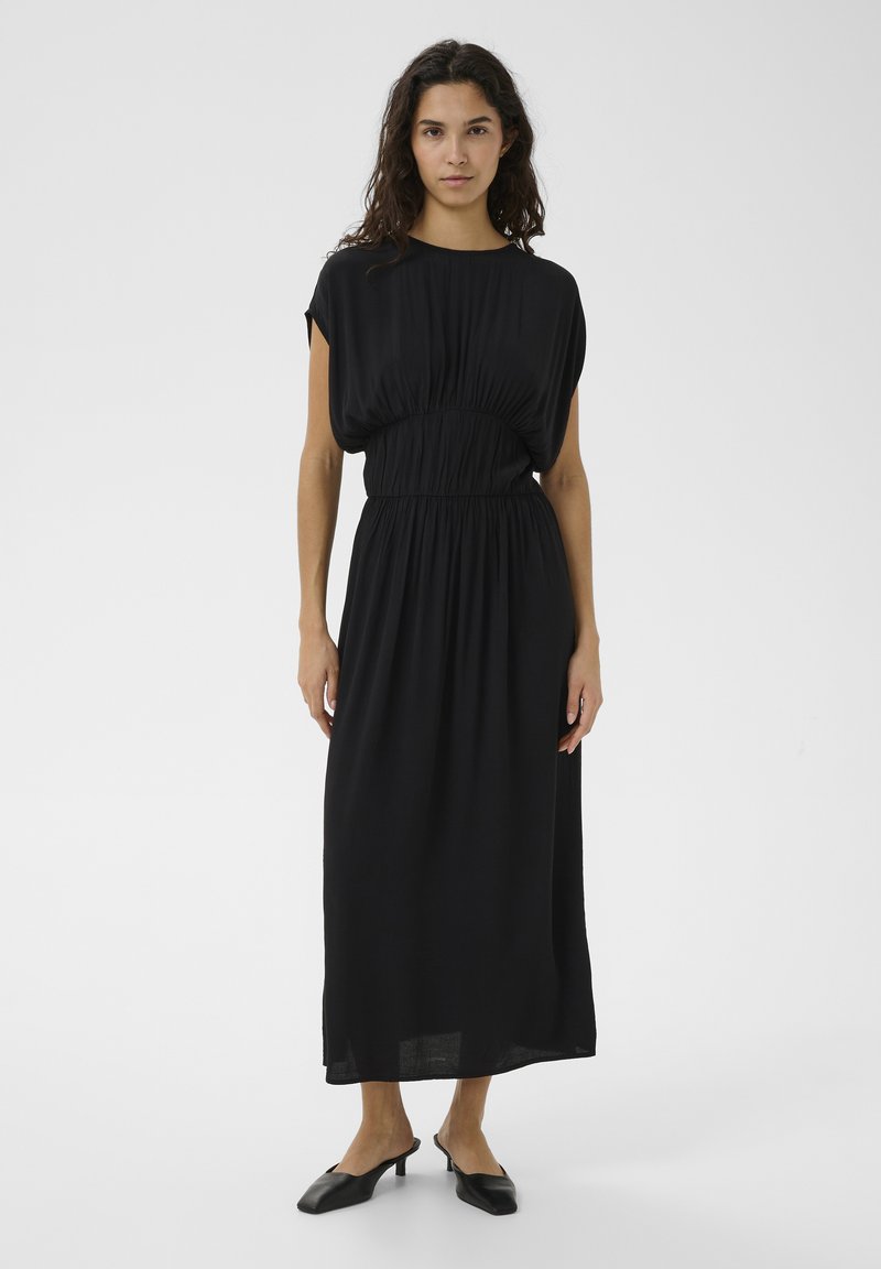 Femme aux longs cheveux bouclés portant une robe midi noire ample à taille froncée et des mules à petit talon noir, debout sur un fond blanc.
