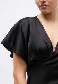 Μαύρο φόρεμα από σατέν με βαθύ V-λαιμόκοψη, μανίκια που κυματίζουν και εφαρμοστό μπούστο. Ομαλή υφή και κομψό draping αναδεικνύουν το σχέδιο.