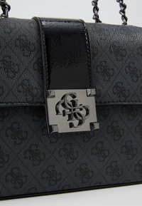 Sac à main design noir avec motif de logo en relief, sangle noire brillante et fermoir métallique argenté avec des lettres entrelacées.