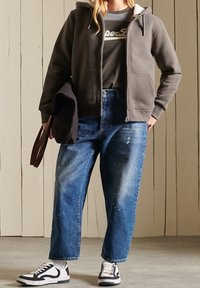 Sudadera gris con cremallera sobre una camiseta oscura, combinada con jeans de pierna ancha desgastados de color azul claro y zapatillas negras y blancas. Bolsa marrón sujetada en la mano.