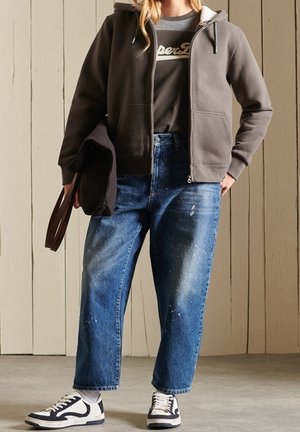 Sudadera gris con cremallera sobre una camiseta oscura, combinada con jeans de pierna ancha desgastados de color azul claro y zapatillas negras y blancas. Bolsa marrón sujetada en la mano.