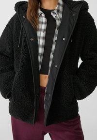 Schwarze Kunstfelljacke mit Reißverschluss und Druckknopfverschluss, ausgestattet mit einer Kapuze, strukturierter Oberfläche und einer kontrastierenden Innenauskleidung mit einem grauen Karomuster.