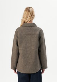 Blond persoon met zijn rug naar de camera in een taupe textuur fleece jacket met lange mouwen en donkere broek, tegen een witte achtergrond.