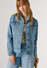 Kvinde iført lyseblå denimjakke og jeans over en hvid ribbet tanktop mod en ensfarvet baggrund.