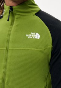 Zielona kurtka zapinana na zamek błyskawiczny z czarnymi rękawami, z teksturowaną powierzchnią i logo "The North Face" na lewej piersi.