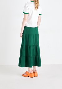 Valkoinen lyhythihainen paita, jossa on vihreät yksityiskohdat, yhdistettynä pitkään vihreään kerrokselliseen hameeseen ja oransseihin platform-sandaaleihin. Silkkinen pinta, kerroksellinen muotoilu.