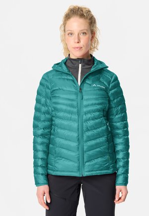 Frau mit hellem lockigem Haar, die eine türkisfarbene gesteppte Jacke mit Kapuze und schwarze Hose trägt, blickt nach vorne vor schlichtem Hintergrund.