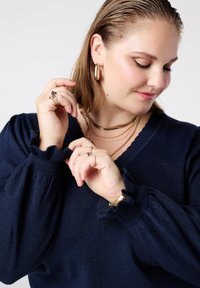 marineblauwe gebreide trui met geplooide manchetten, V-hals en een zachte textuur. Model draagt meerdere gouden kettingen en ringen, met aandacht voor de sieradendetails.