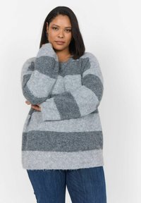 Pull rayé gris et gris clair, fabriqué en matière douce et moelleuse. Col rond, épaules tombantes et coupe décontractée. Associé à un jean en denim foncé.