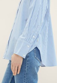 Camisa a riscas azul e branca com ajuste descontraído, punhos com botão e barra arredondada, combinada com jeans de ganga azul claro.