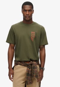 Camiseta de algodón verde oliva de manga corta, con texto y logo rojos en vertical. Combinada con pantalones beiges y una camisa de cuadros atada a la cintura.