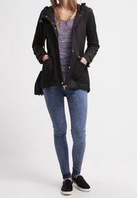 Black hooded jacket with zip closure and two front pockets, paired with a patterned purple top, blue denim-like leggings, and black sneakers.
