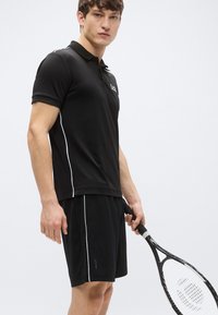 Čierna polokošeľa s rebrovaným golierom a bielymi bočnými akcentmi, kombinovaná s čiernymi krátkymi nohavicami. Osoba drží čiernu tenisovú raketu.