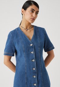 Vestido de ganga com decote em V, mangas curtas e fecho de seis botões. Apresenta costuras verticais e uma textura suave. Na cor azul média.