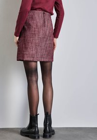 Falda mini de tweed color burdeos con cierre de cremallera en la parte posterior, combinada con medias opacas negras y botas de tobillo sobre un suelo gris.