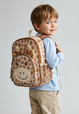 Young boy wearing light blue shirt and beige pants, looking back, carrying a giraffe-patterned backpack with plush ears and antennae.