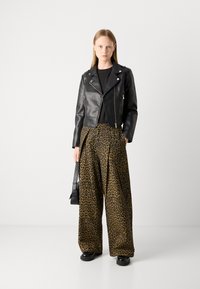 Veste en cuir noire coupée, chemise noire et pantalons larges à imprimé léopard. Le modèle se tient avec un petit sac noir et des chaussures noires épaisses.