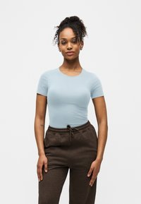 Νεαρή γυναίκα με σγουρά μαλλιά, φορώντας ανοιχτό μπλε στενή μπλούζα και σκούρο καφέ παντελόνι με κορδόνι, στέκεται μπροστά σε λευκό φόντο.