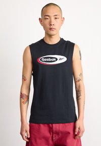 Juodas be rankovių medvilninis mar�škinėliai su centrinio balto Reebok logotipo raudonu kontūru, dėvimi su raudonomis šortais, matomi tatuiruotės ant rankų.