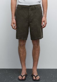 Dark green cotton shorts with a straight cut, flat front, and belt loops. Worn with black flip-flops, features minimal stitching and a button closure.