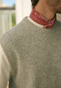 Szary sweter z dzianiny o teksturowanym wzorze, z widoczną białą koszulką pod szyją. Na szyi związana jest czerwona bandana w biały wzór paisley.