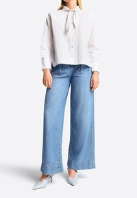 Mujer vistiendo una blusa blanca de manga larga con un lazo en el cuello, jeans de pierna ancha en azul claro y tacones de punta en azul claro.