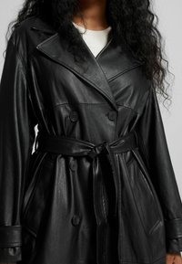 Trench coat de cuero negro con cierre de botones de doble botonadura y un lazo en la cintura. Presenta amplios solapas y bolsillos laterales, con un acabado texturizado.