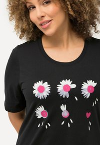 Camiseta negra de manga corta con un estampado floral de margaritas blancas con centros y acentos en rosa, diseñada con un cuello redondo.