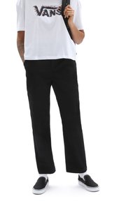 Sorte bukser med lige pasform, parret med en løs hvid t-shirt med et flerfarvet "VANS" logo og sorte slip-on sko.