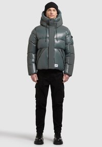Kurtka puchowa w błyszczącym ciemnoszarym kolorze z wysokim kołnierzem, zamkiem błyskawicznym i regulowanymi sznurkami. Połączona z czarnymi spodniami typu cargo i butami.