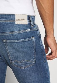 Jeans de mezclilla azul para hombre con un lavado claro, que presentan un bolsillo trasero con un patrón de costura en zigzag y una etiqueta de marca de cuero posicionada arriba.