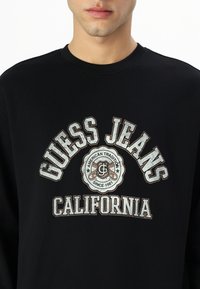 Sudadera negra de manga larga con el logo "GUESS JEANS CALIFORNIA" en gris y blanco, con un emblema circular en el centro.