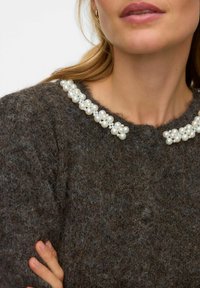 Tumši brūns džemperis ar pūkainu faktūru, rotāts ar dekoratīvu apkakli, kas izgatavota no baltām pērļu krellēm.
