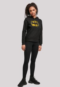 Black hoodie with a yellow Batman logo, featuring a front pouch pocket and drawstrings, paired with black leggings and sleek shoes.