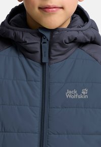 Blå vadderad jacka med hög krage, dragkedja och kontrasterande mörkblå axelpanel. Har logotyp på bröstet i ljusgrå.