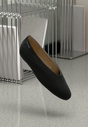 Scarpa piatta nera slip-on di Calvin Klein fluttua accanto a un supporto metallico con borse della spesa bianche su un pavimento grigio.