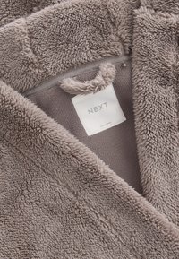 Tissu doux et duveteux de couleur taupe avec une étiquette de vêtement blanche cousue portant l'inscription "NEXT" et "Fabriqué en Chine".