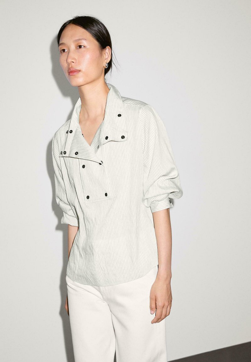 Lys grå stribet skjorte med kravesnit, knapdetaljer og løse ærmer, parret med hvide bukser i et minimalistisk miljø.