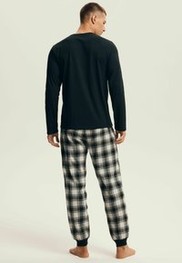 Mężczyzna stojący boso, ubrany w czarną koszulę z długim rękawem oraz czarno-białe, kraciaste spodnie piżamowe, zwrócony tyłem na jednolitym tle.