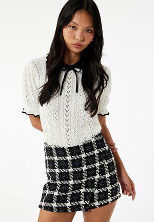 Witte gebreide blouse met korte pofmouwen en een zwarte scalloped kraag. Gecombineerd met een zwart-wit geruite minirok.