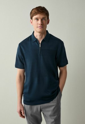 Dunkelblaues Polohemd mit kurzem Reißverschluss und strukturierten horizontalen Streifen, kombiniert mit hellgrauen Hosen. Glatter Stoff, kurze Ärmel.