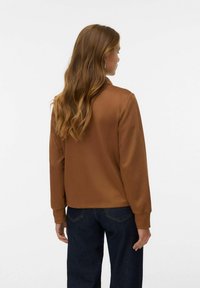 Braune Satinjacke mit Kragen und langen Ärmeln, ausgestattet mit gerippten Bündchen. Getragen mit dunklen Jeans, die eine glatte Textur und einen maßgeschneiderten Sitz zeigen.