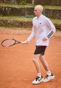 Jachetă atletică albă cu dungi negre, pantaloni scurți negri și pantofi de tenis. Ținând o rachetă de tenis, pe un teren de zgură cu un fundal stâncos.