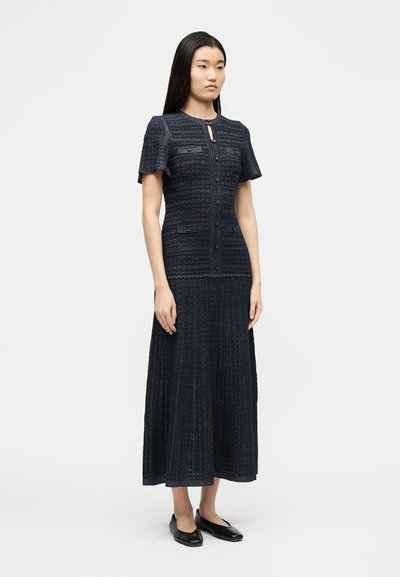 Γυναίκα που στέκεται φορώντας μπλε σκούρα υφ ασμένη κοντομάνικη μπλούζα και ταιριαστή μακριά φούστα με μαύρες μπαλαρίνες σε απλό λευκό φόντο.