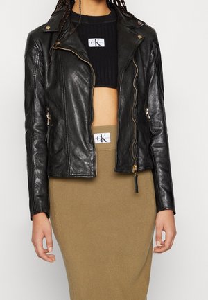 Žena nosící černý krátký svetr, černou koženou bundu se zipem a béžovou žebrovanou sukni s vysokým pasem s viditelnými logy Calvin Klein.