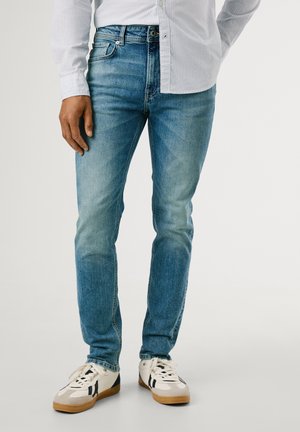 Jeans de denim azul con silueta ajustada, que presentan detalles desgastados, pasadores para cinturón y diseño de cinco bolsillos, complementados con zapatillas blancas.
