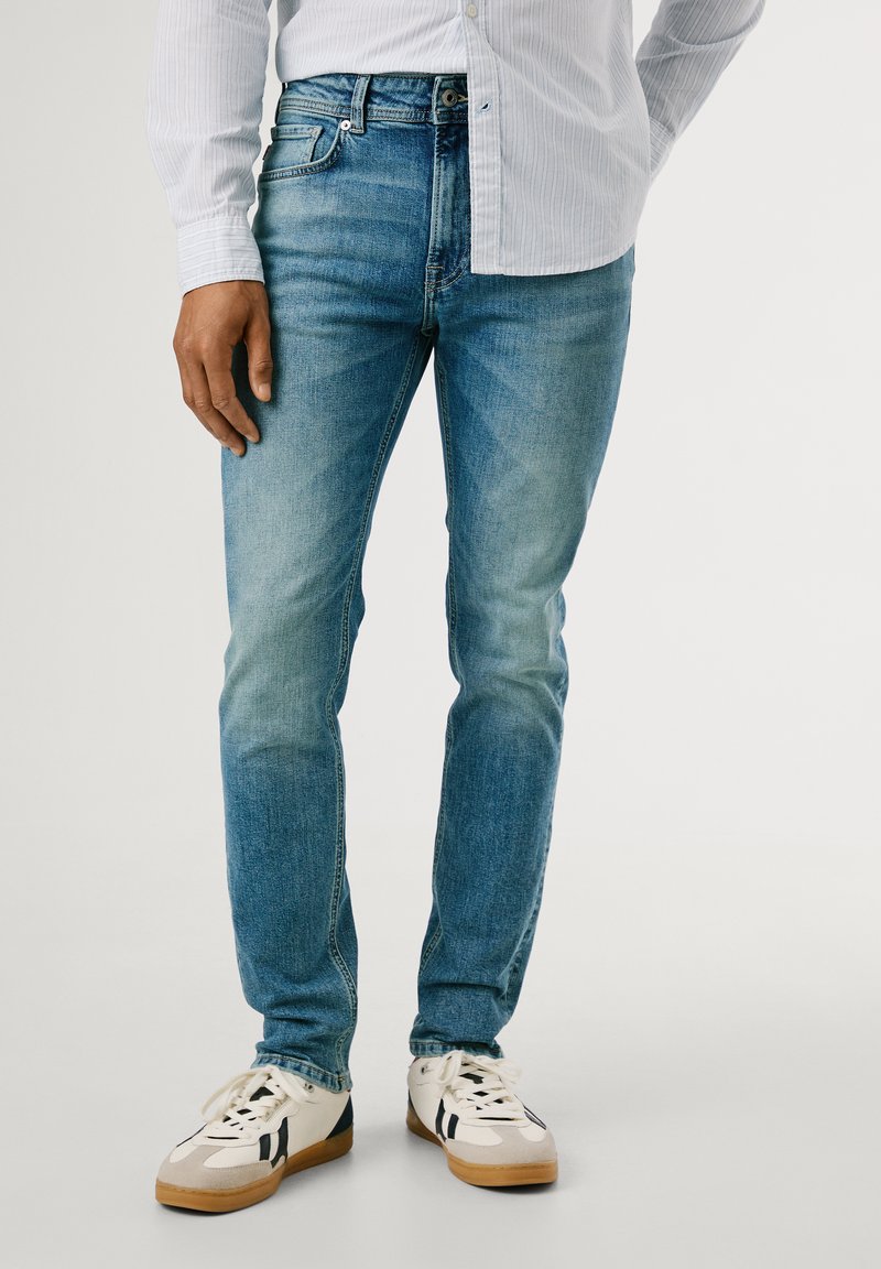 Jeans de denim azul con silueta ajustada, que presentan detalles desgastados, pasadores para cinturón y diseño de cinco bolsillos, complementados con zapatillas blancas.