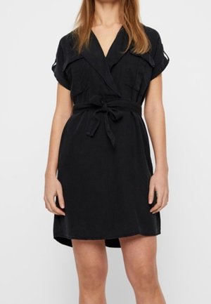 Kobieta ubrana w czarną sukienkę na krótki rękaw z wiązaniem w talii i kieszeniami na piersi, stojąca na tle jednolitego białego tła.
